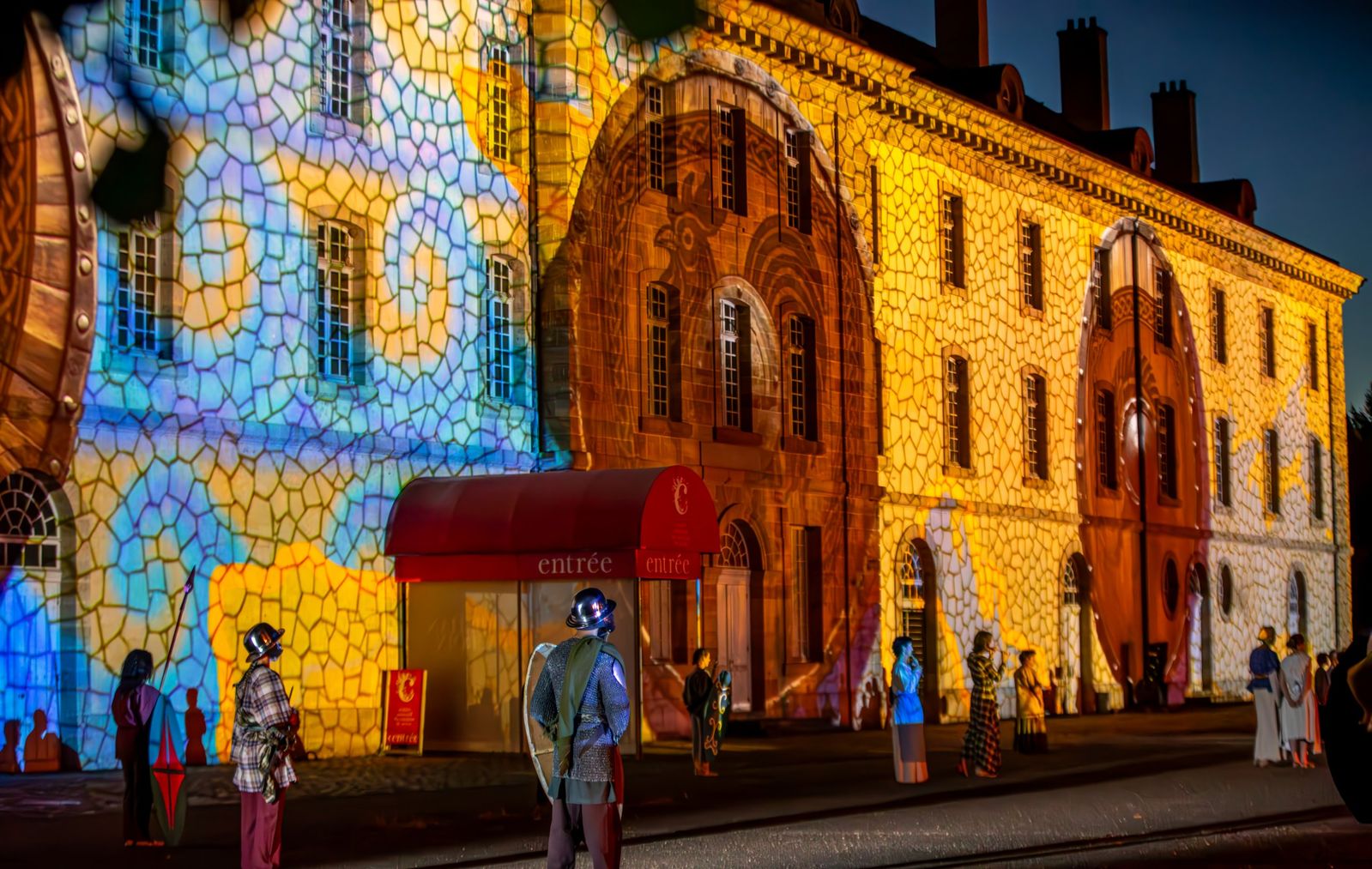 Une façade aux mosaïques de couleurs projetées, avec des figurants en costume