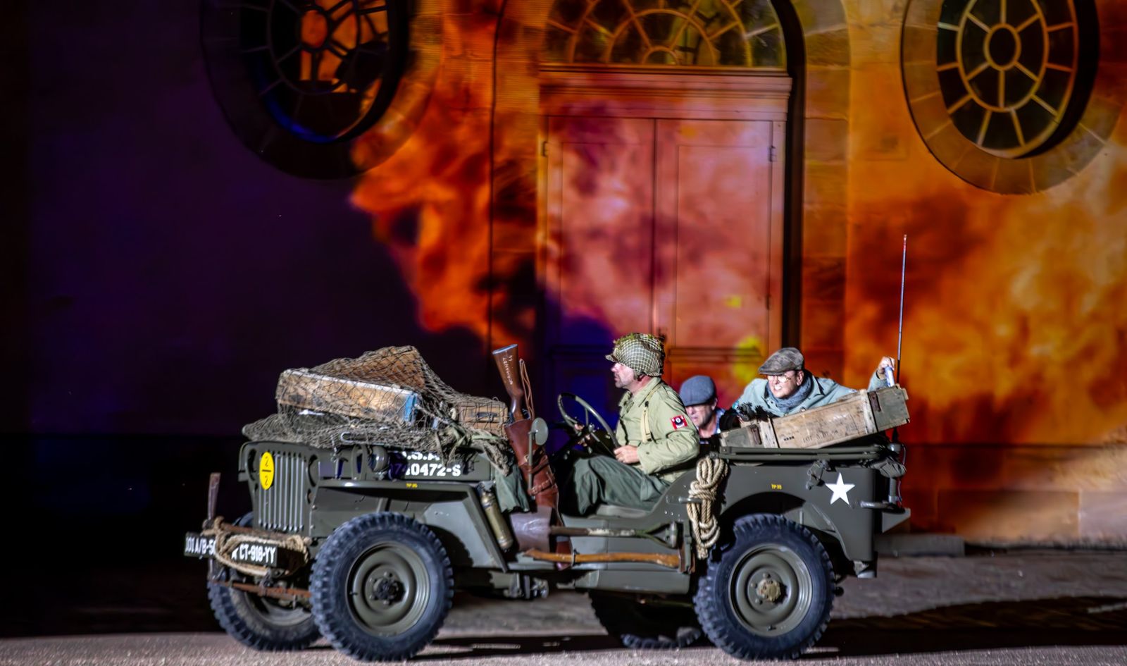 Une jeep militaire d’époque devant une façade embrasée de flammes projetées